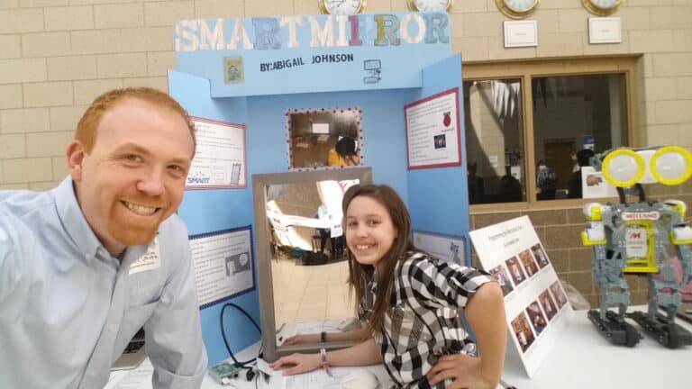 Innovative technology project display at school science fair featuring a girl and adult smiling with a robotic figure and creative posters.