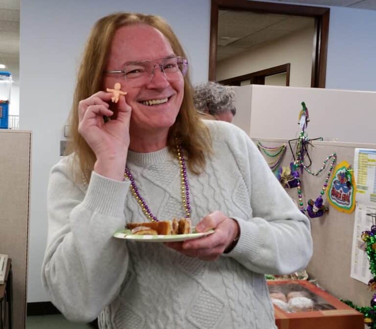 Festive office celebration with cheerful woman holding holiday treats and wearing Mardi Gras beads at Twin State Technical Services.
