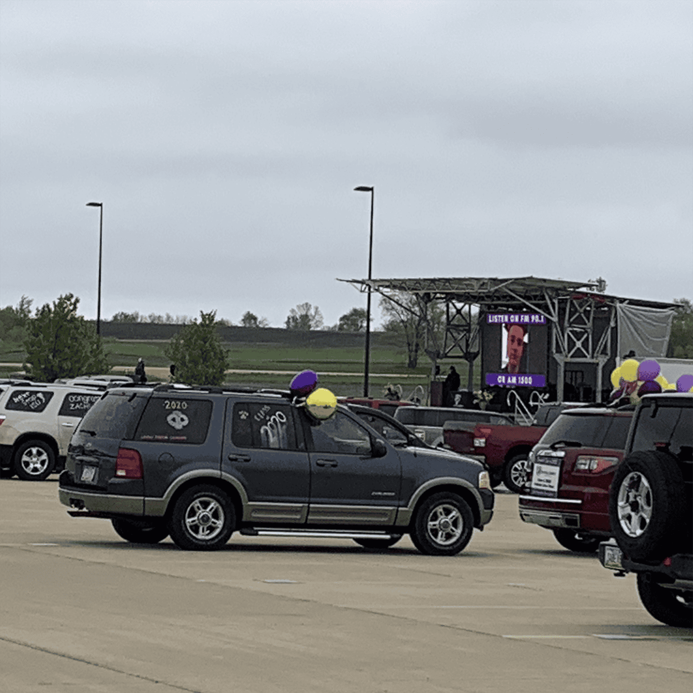 Roadside event with parking lot, vehicles decorated with balloons, outdoor stage, and a live music performance, promoting community engagement and local events.